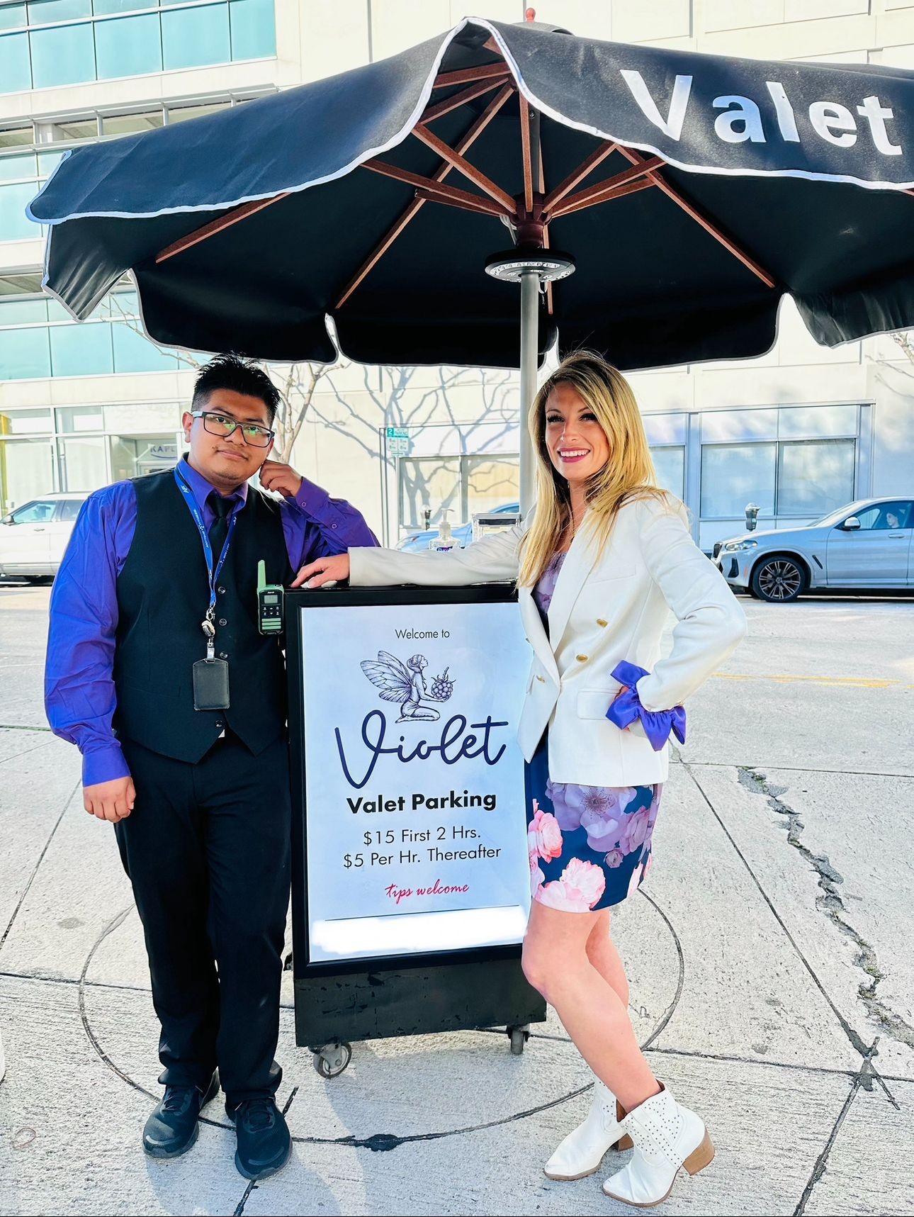 Two people standing next to a valet parking sign under a black umbrella outside a building.