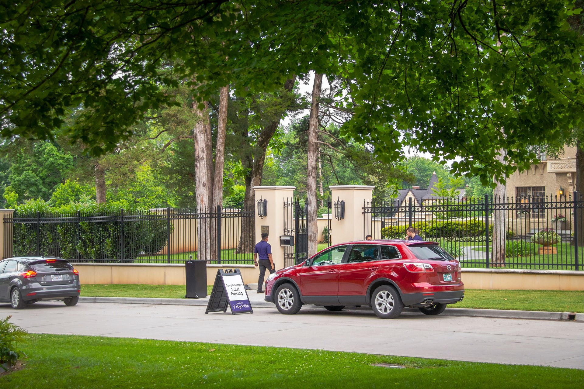 Valet parking on neighborhood street outside luxious fenced estate with many tall trees.